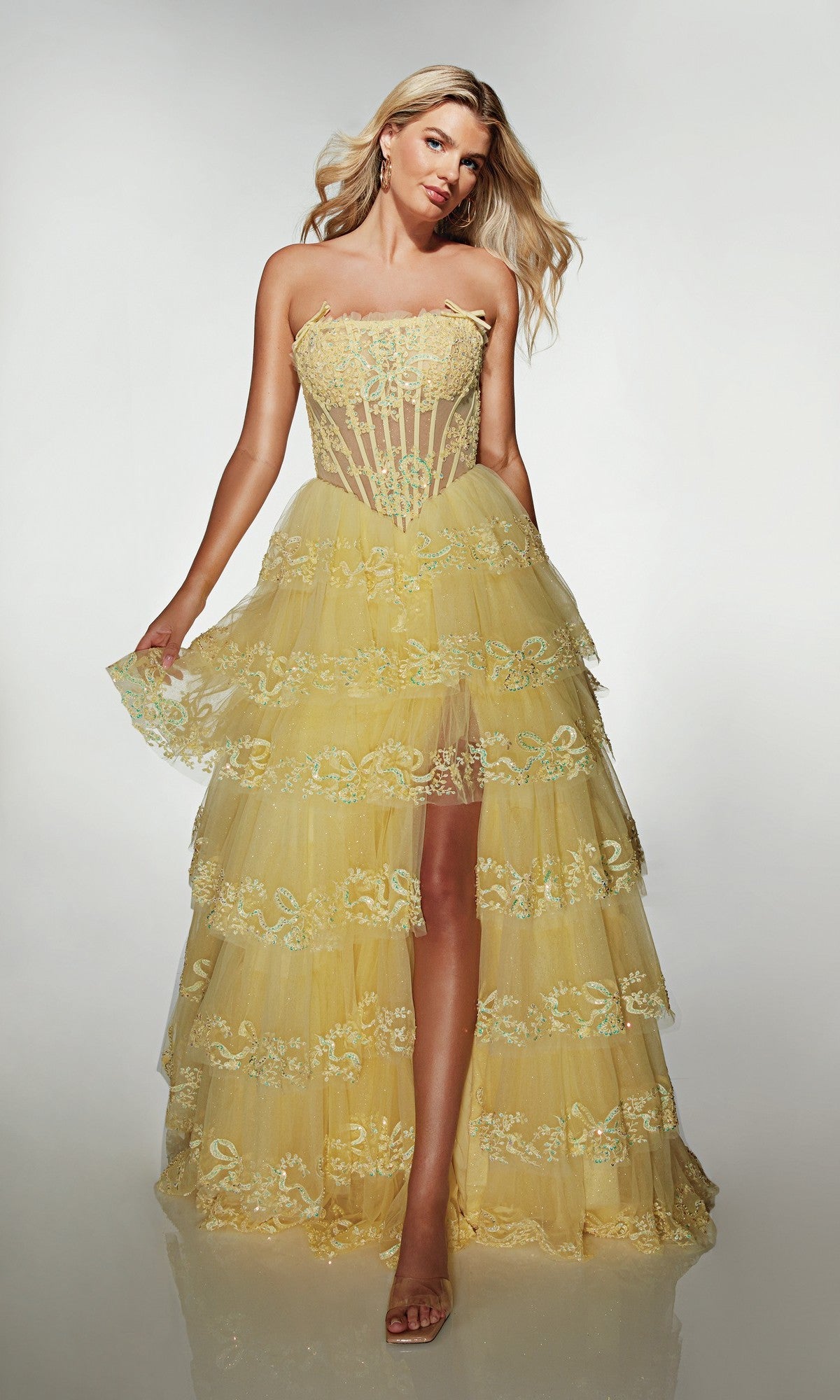 A woman models the Alyce 62191 long prom dress in light yellow, featuring a strapless floral-embroidered sheer corset bodice and tiered tulle A-line skirt, posing against a plain light background.